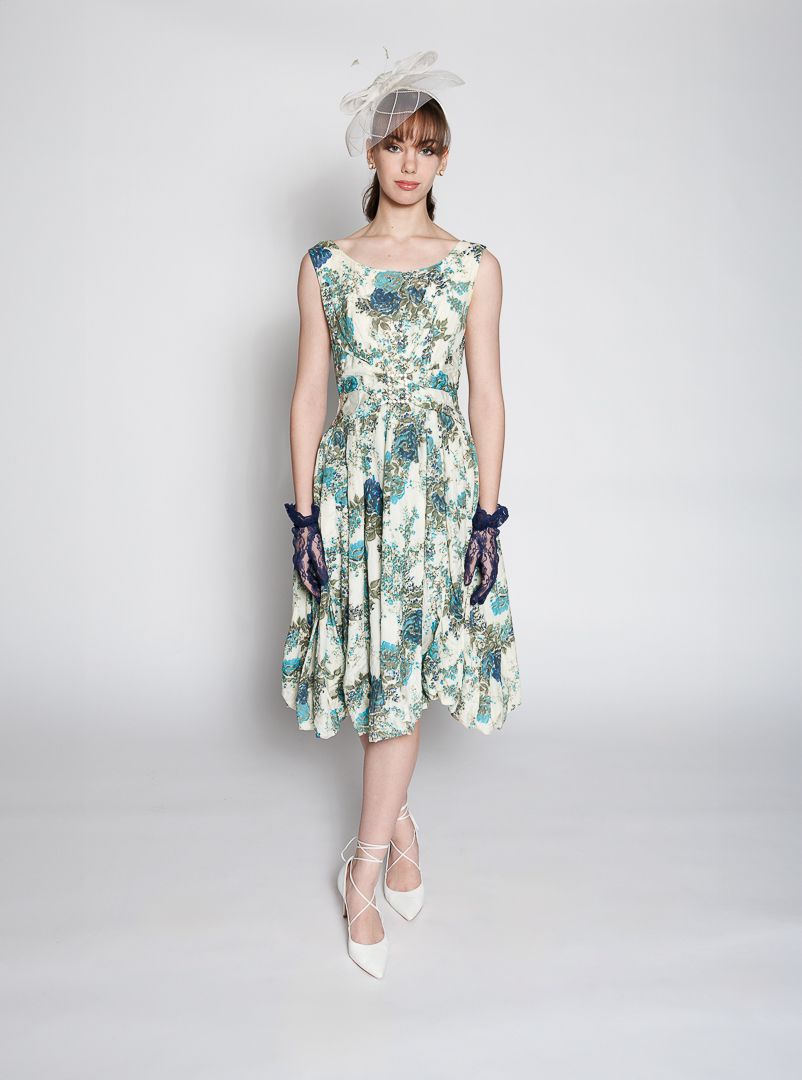 Woman in floral teal dress, white fascinator hat, purple gloves, and white heels posing elegantly.