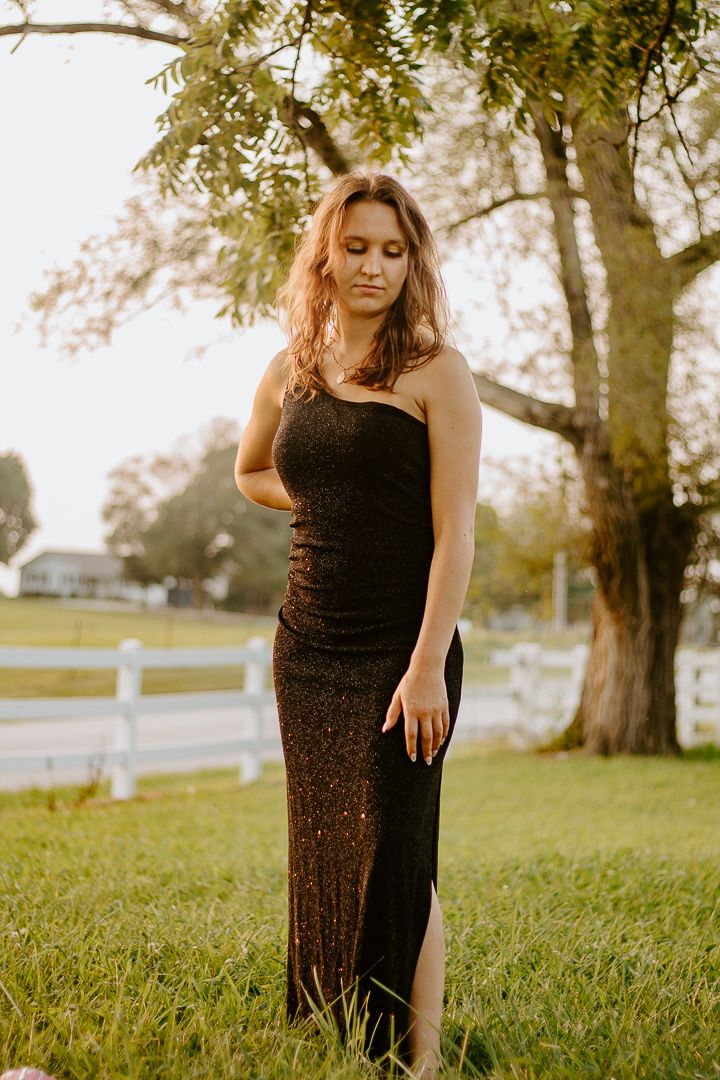 Young woman in a black sequin strapless gown poses outdoors on green grass near a white fence and tree.
