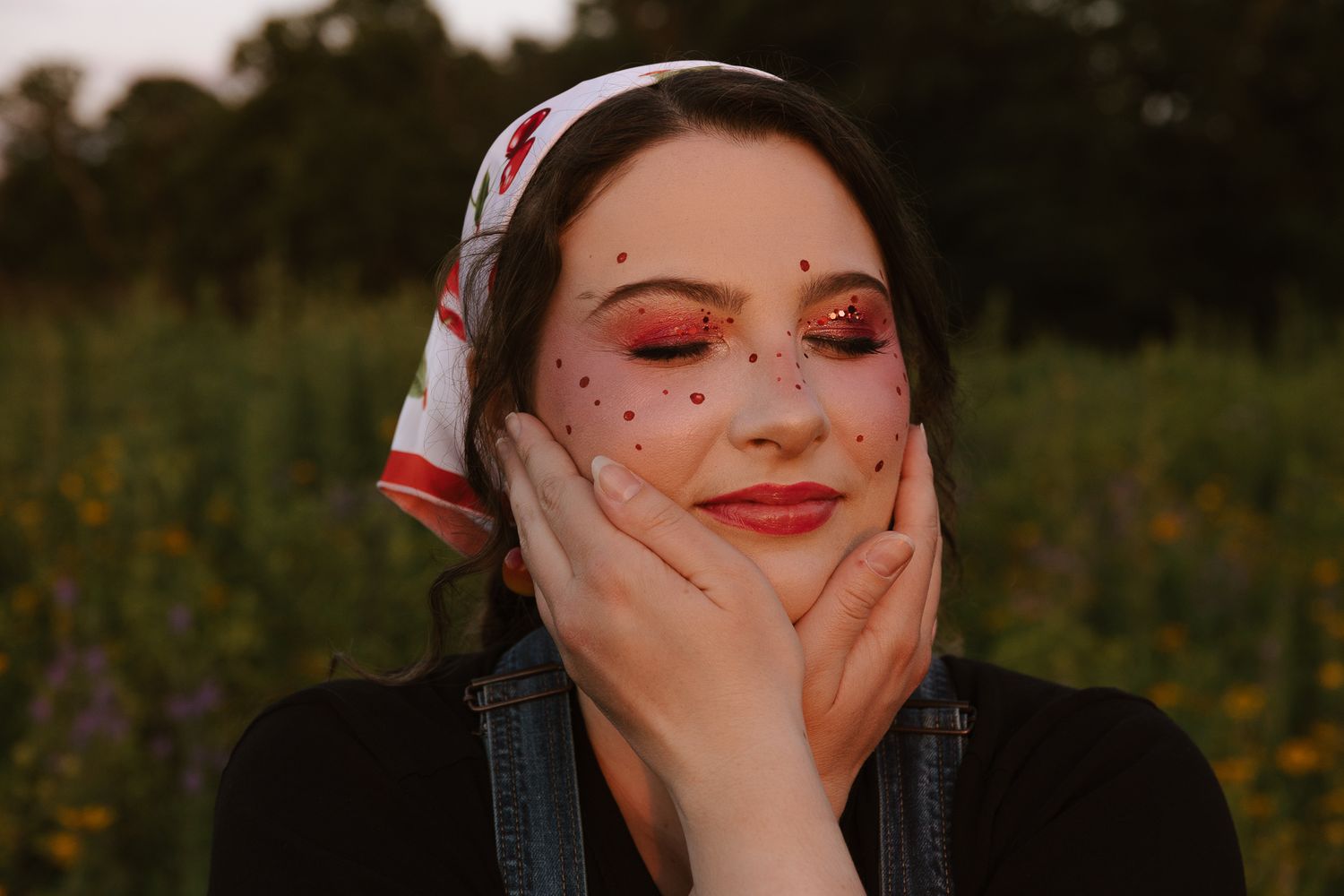 Young woman with polka dot veil, rosy eye makeup, and cherry print headscarf cradling her face in a flower field.