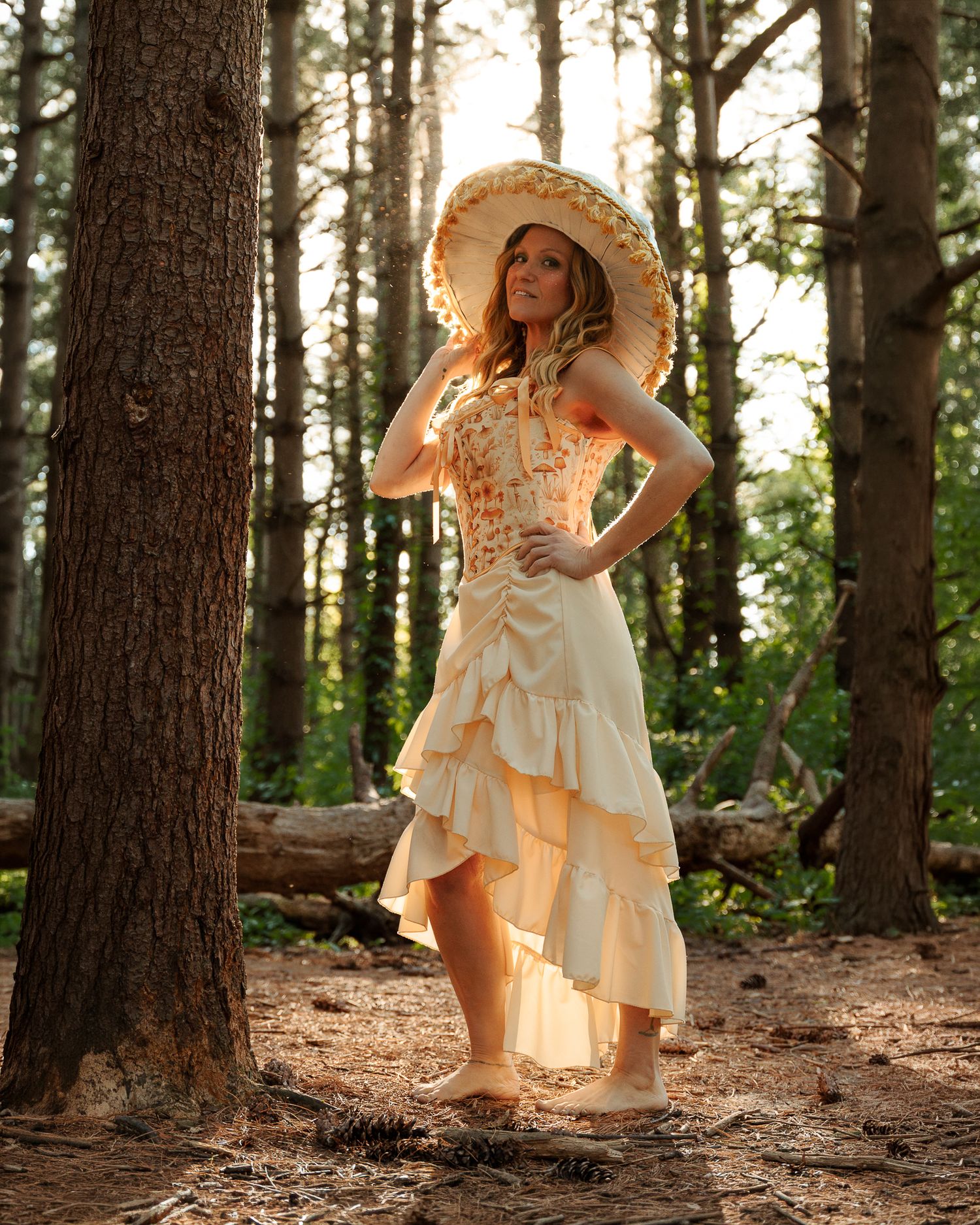 Woman in ruffled cream dress and wide-brim hat poses gracefully among tall pine trees in a sunlit forest.