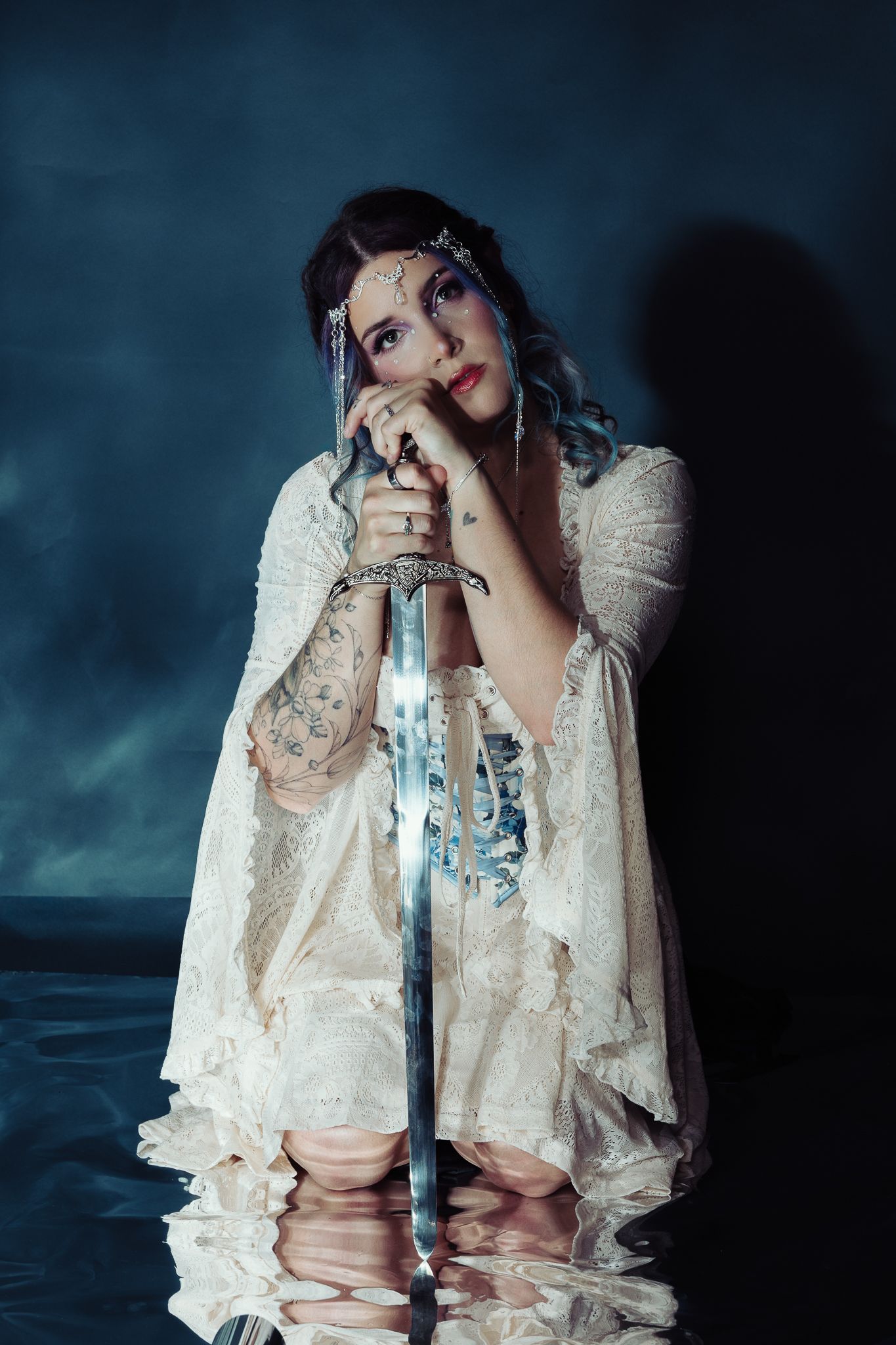 A mystical woman in white lace gown holds a sword, adorned with face gems and tattoos, kneeling in water.