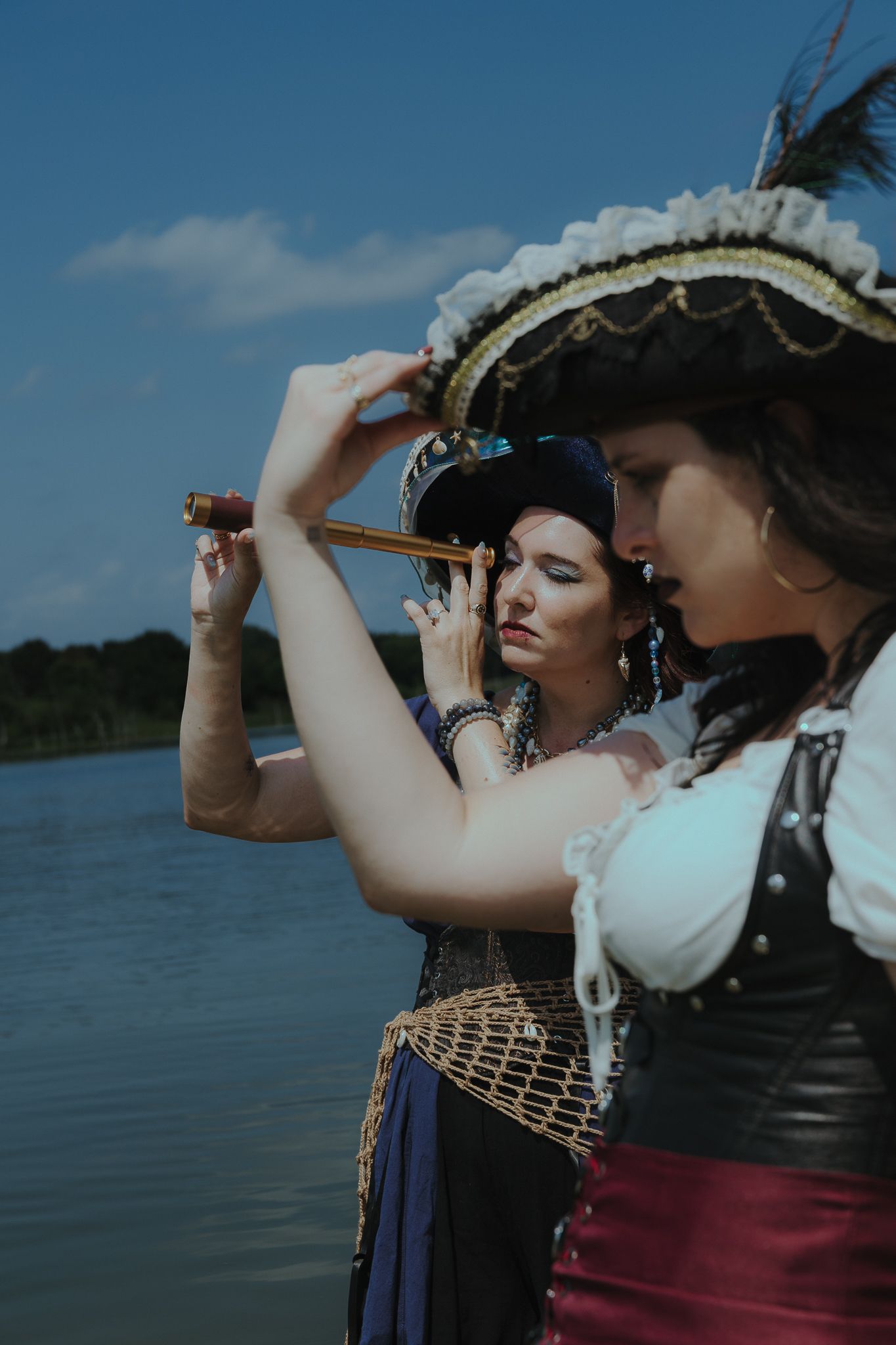 Two women in pirate costumes stand by a lake, one looking through a small telescope under a blue sky.