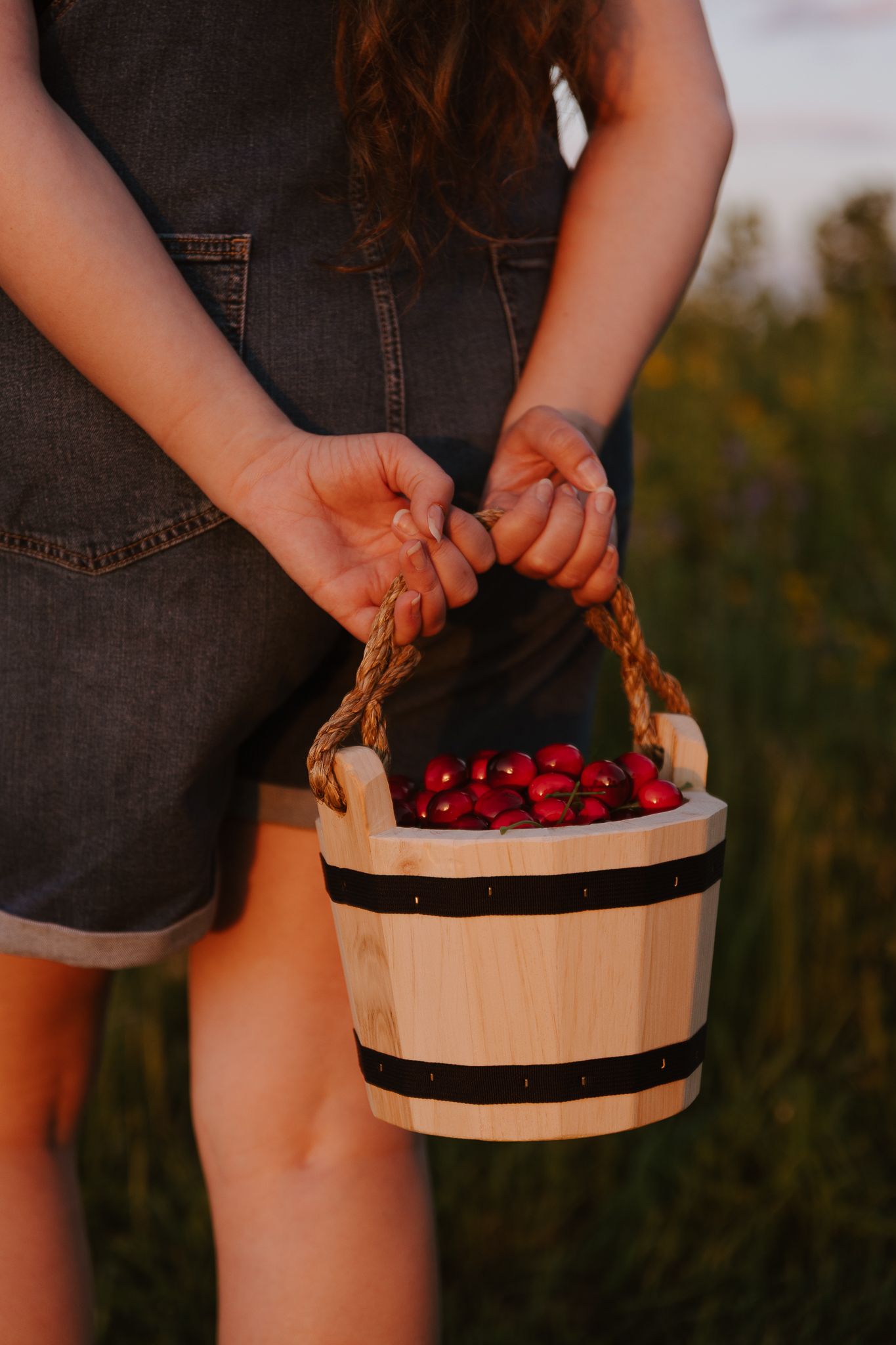 Woman in denim overalls holds a rustic wooden bucket filled with fresh red cherries outdoors.