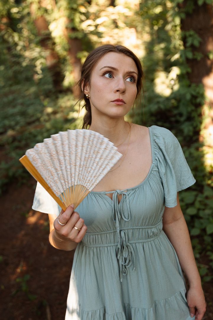 A woman in a sage green dress holds a decorative folding fan in a forest setting.