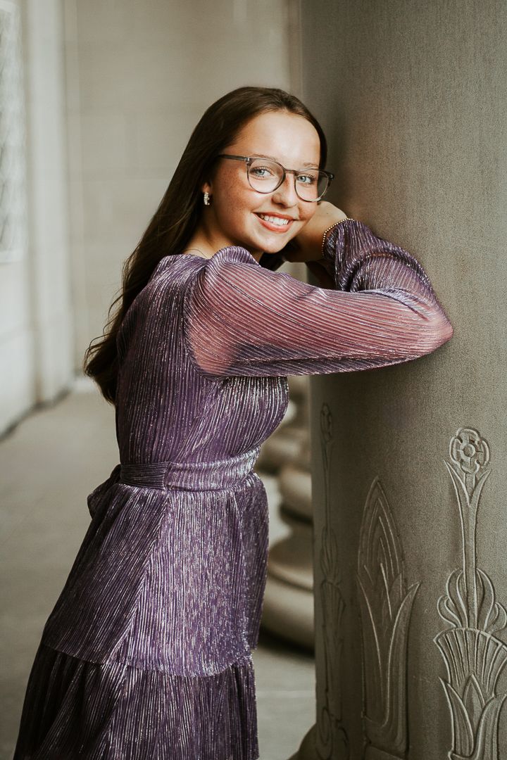 A smiling person wearing glasses poses in a flowing purple metallic dress against a neutral wall.