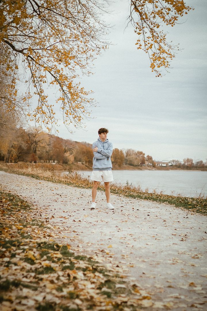 A person stands on an autumn path by a lake with golden leaves and bare tree branches overhead in warm fall lighting.