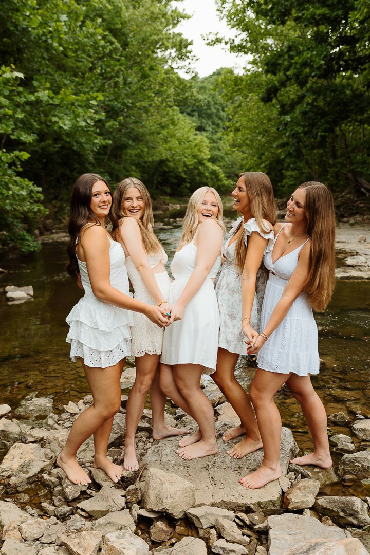 A group in white summer dresses sits together on rocks by a peaceful creek in a wooded setting.