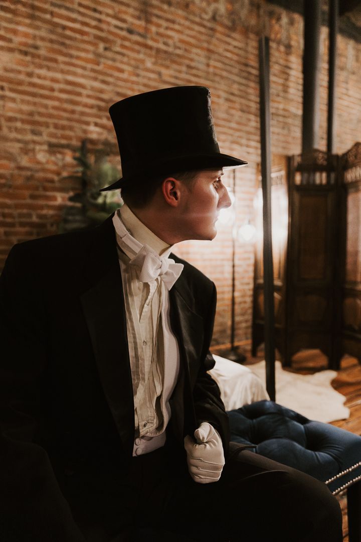 A person wearing a black hat and white shirt sits in profile against a rustic brick wall in moody lighting.
