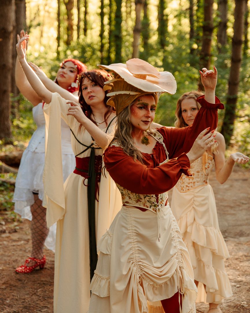 Four women in Renaissance faire costumes dance gracefully in a forest setting, wearing cream and burgundy period dresses.