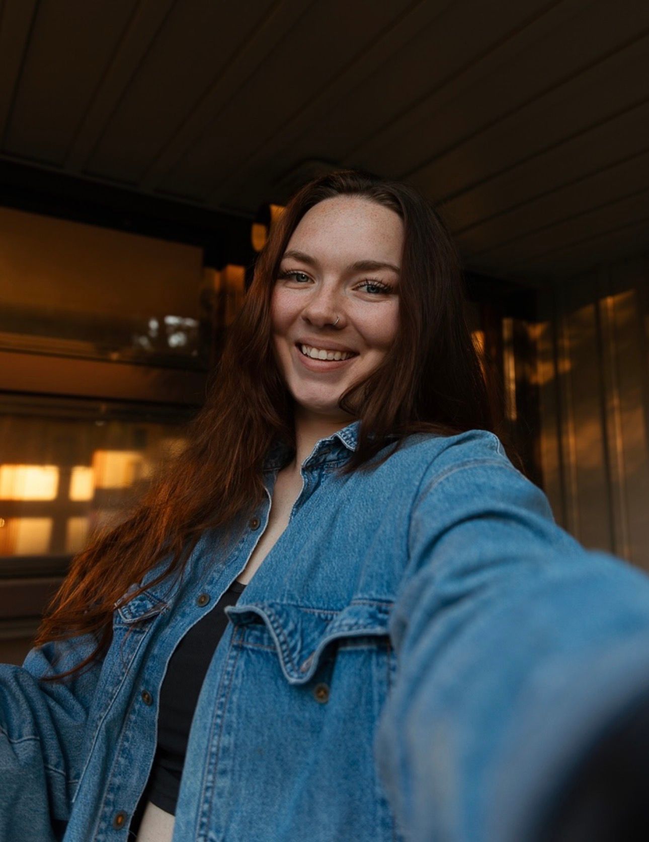 A person wearing a denim jacket poses in a dimly lit indoor setting with warm ambient lighting.