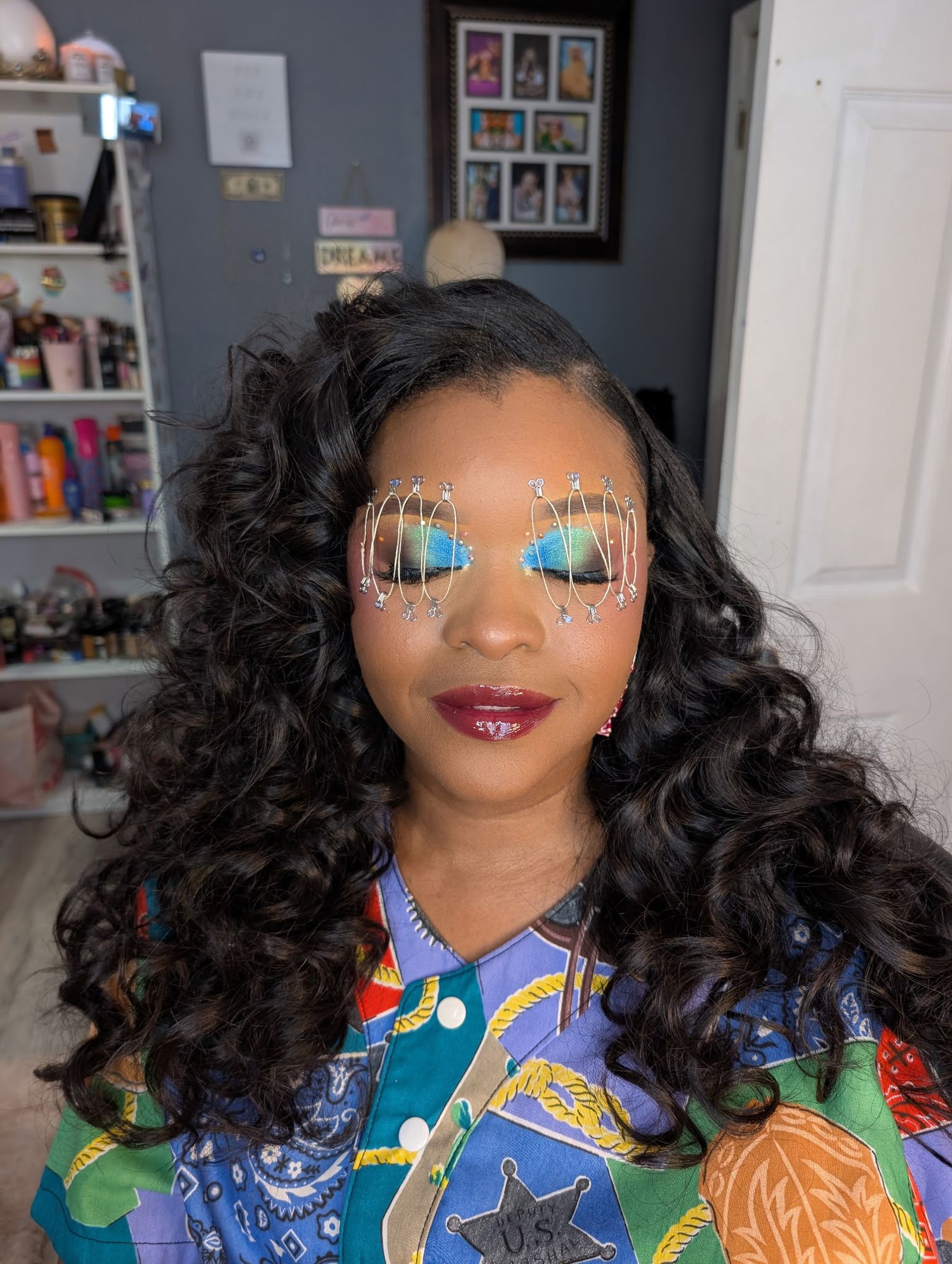 Woman with bold blue eyeshadow, decorative eye gems, dark lips, curly hair, wearing colorful printed shirt.