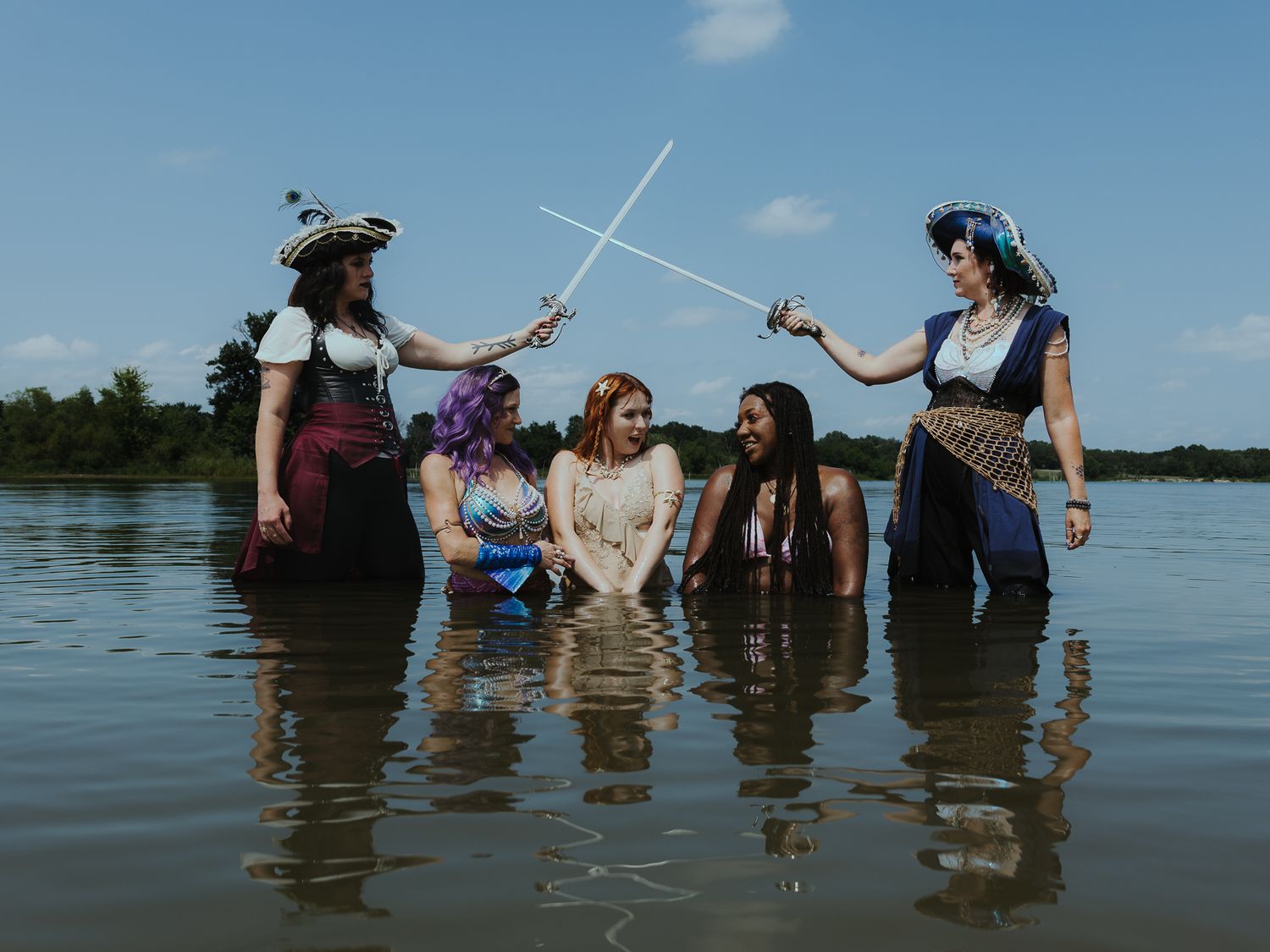 Five women in pirate costumes stand in a lake, two crossing swords while others watch under a blue sky.