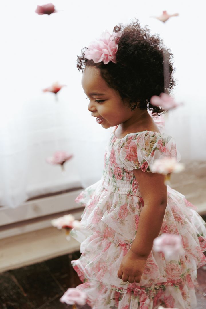 Dreamy portrait of a child in a pink floral dress with floating petals and natural curls adorned with a flower.