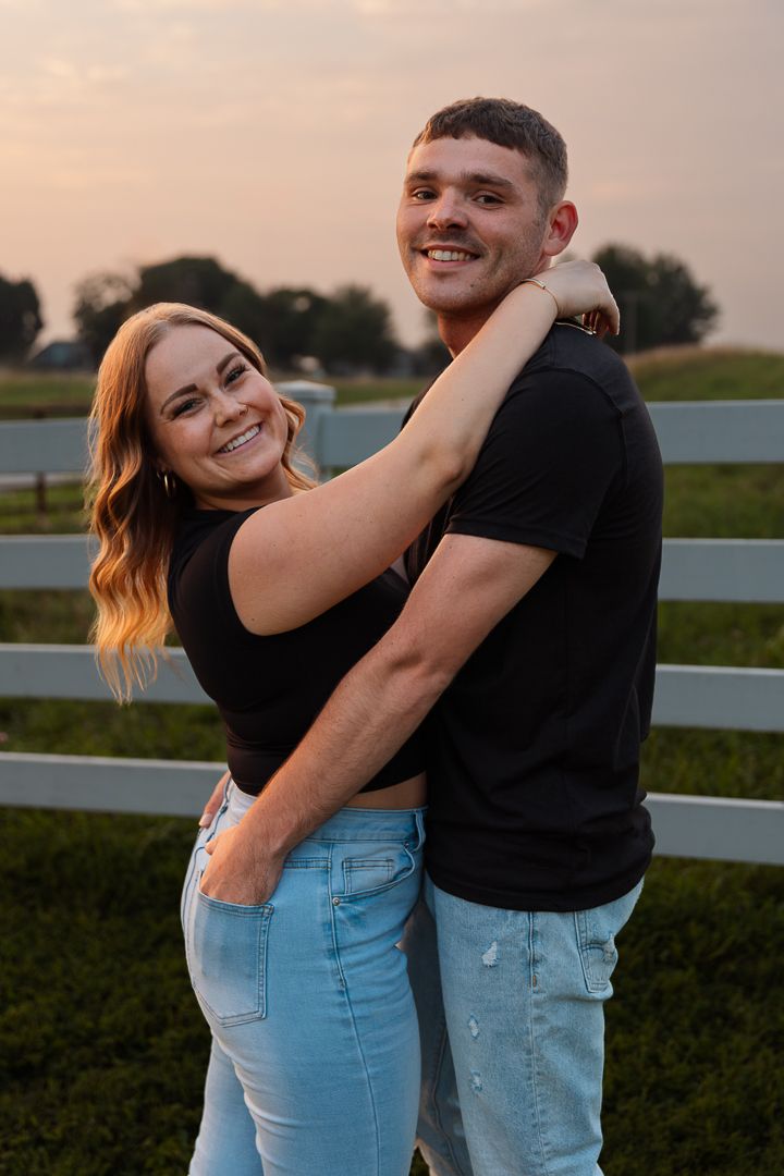 A couple in casual outfits sharing intimate embraces and laughing together against a scenic rural backdrop at sunset.