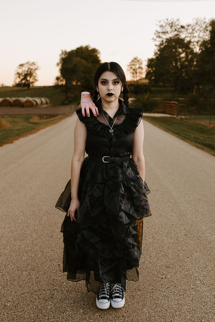 A figure in a dramatic black ruffled dress stands on an empty rural road at sunset.