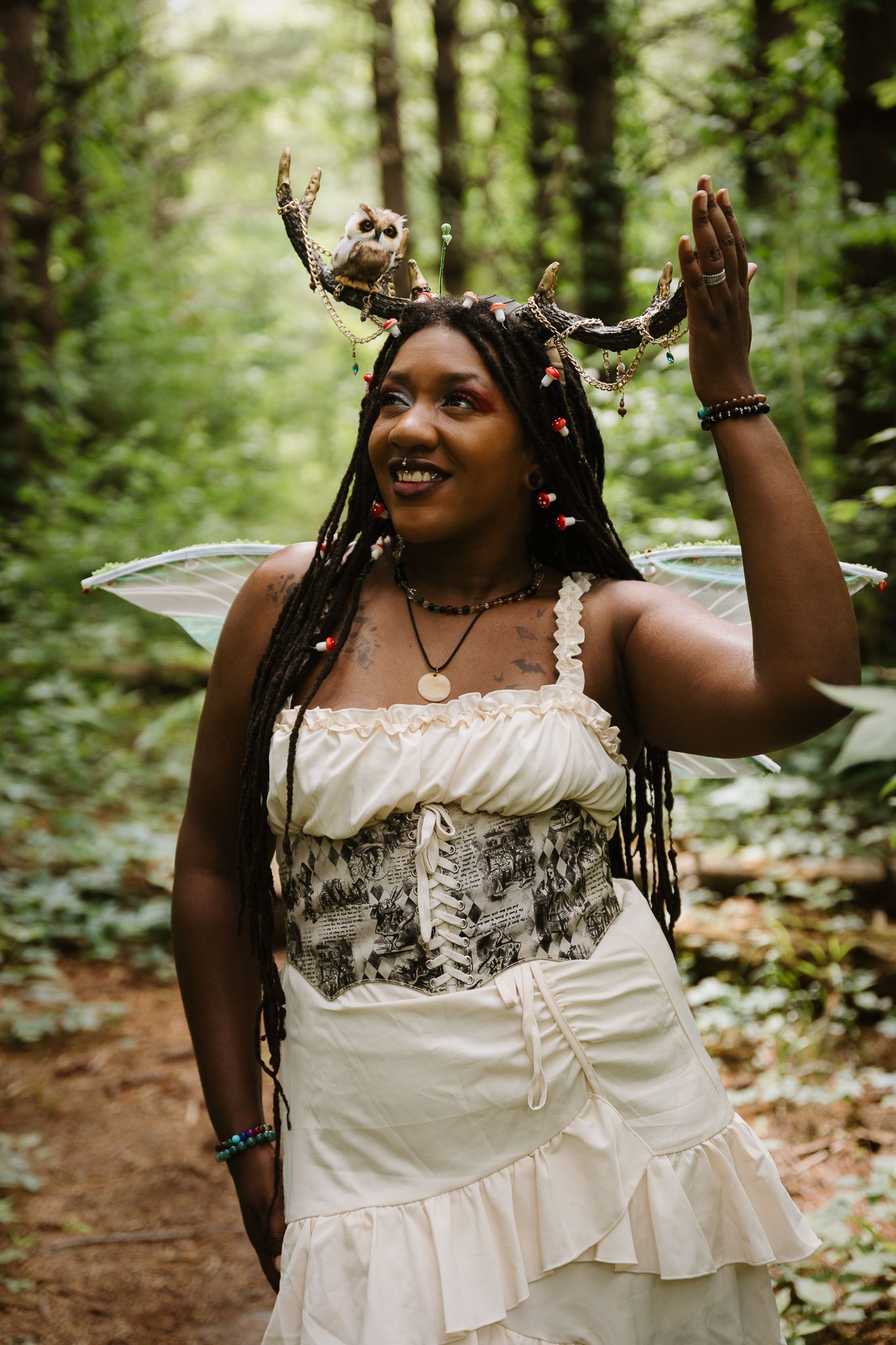 Black woman in fairy costume with owl headpiece and wings poses in lush green forest wearing white dress.