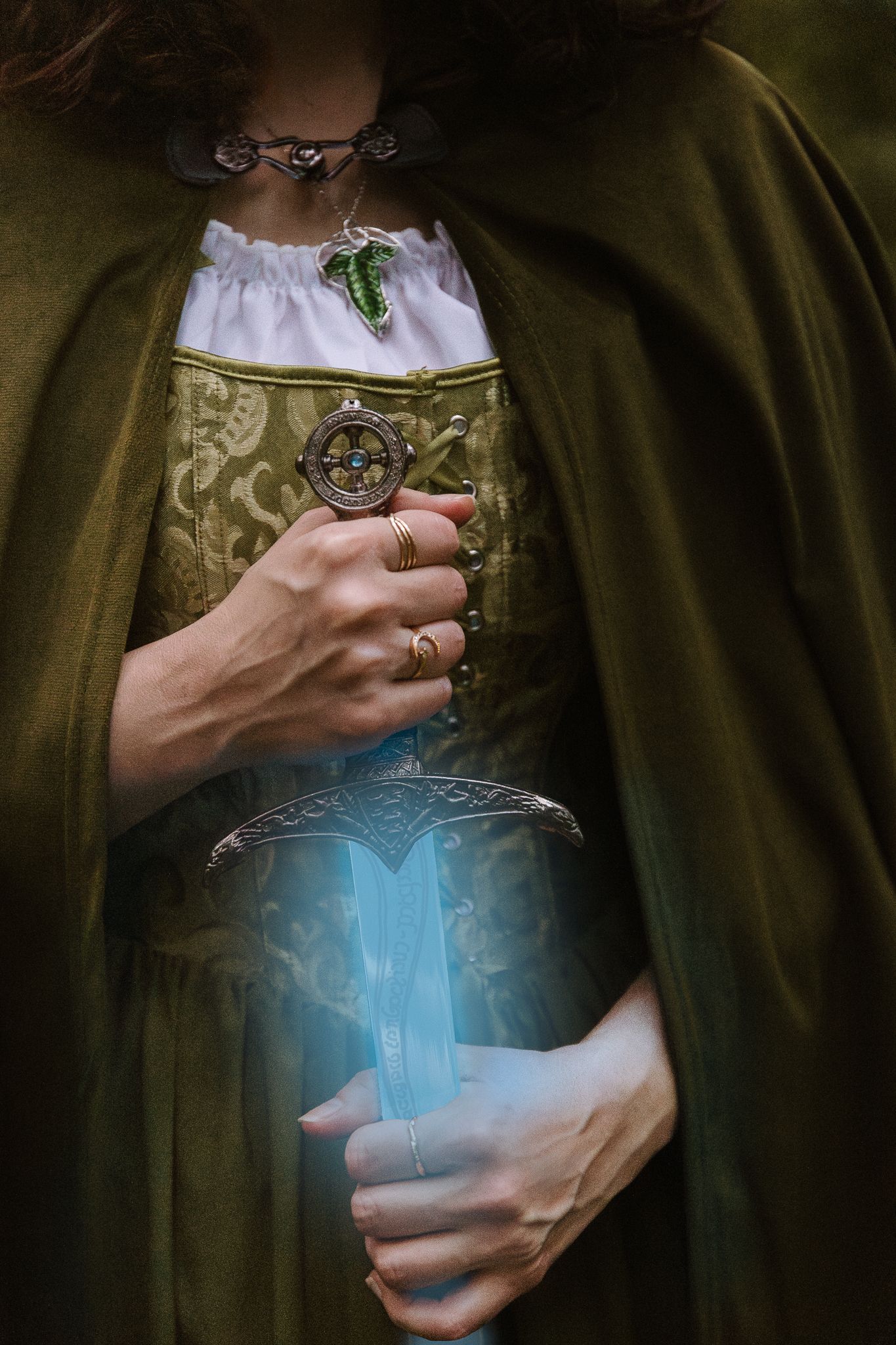 Person in medieval costume holding a glowing magical dagger with ornate jewelry and green cloak.