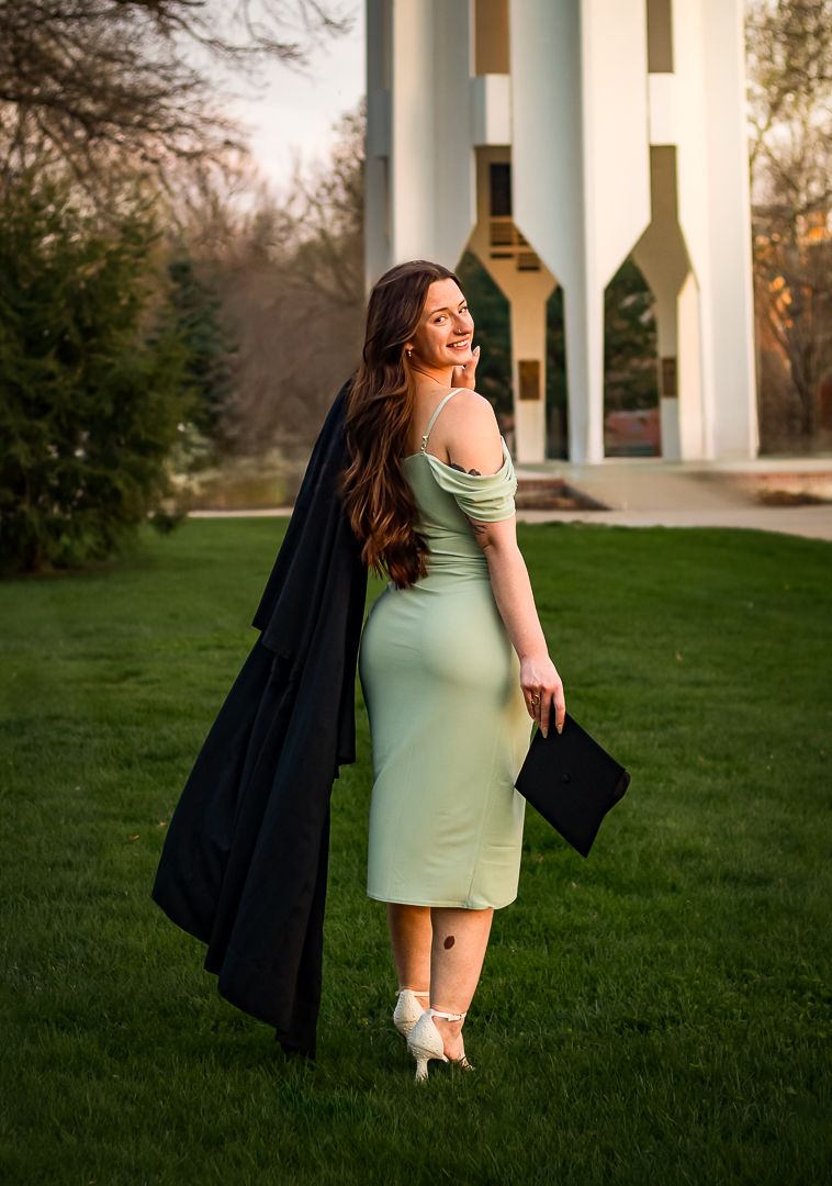 A person in a mint green dress and black jacket poses gracefully on a grassy lawn at sunset.