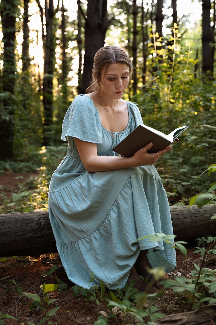 A person in a light blue dress sits on a fallen log in a sunlit forest while reading a book.