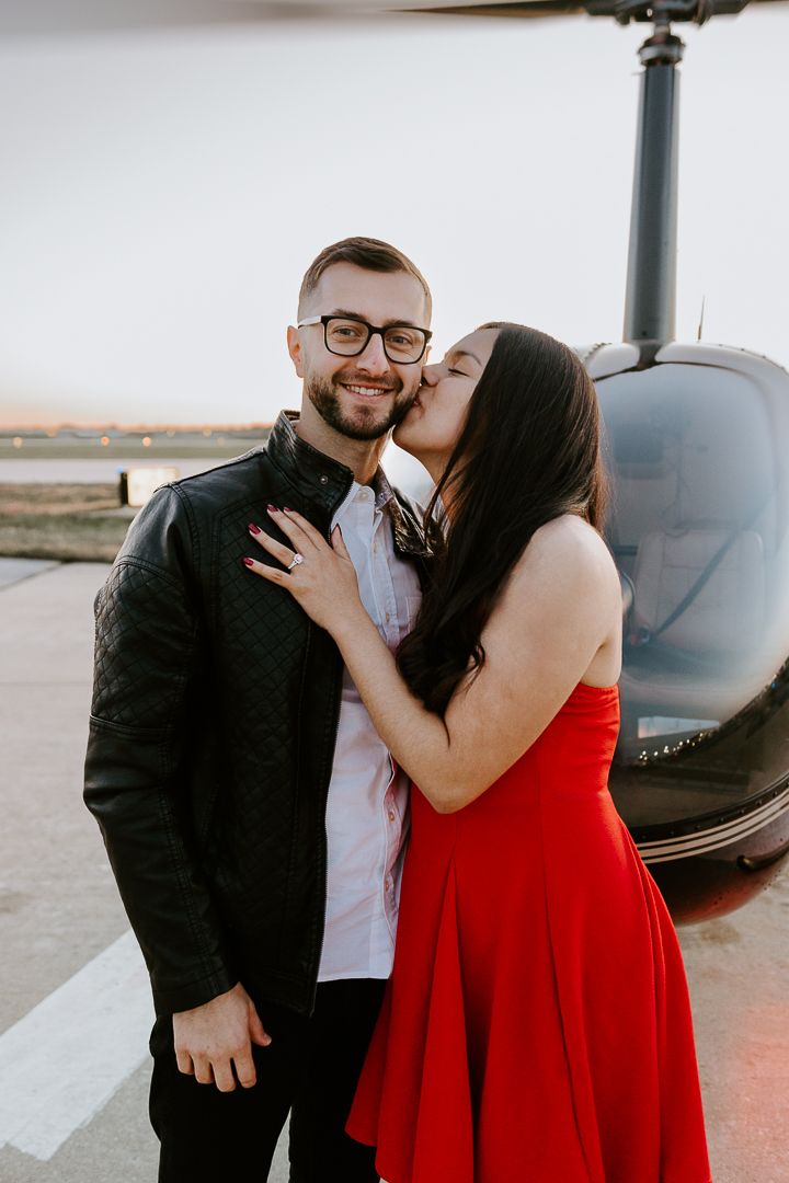 A couple celebrating their engagement next to a parked helicopter at dusk.