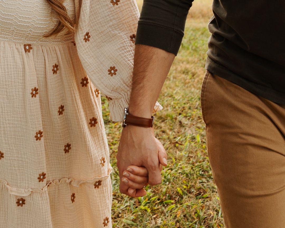 Close-up view of two people holding hands while standing on grass, one wearing white floral dress and other in brown pants.
