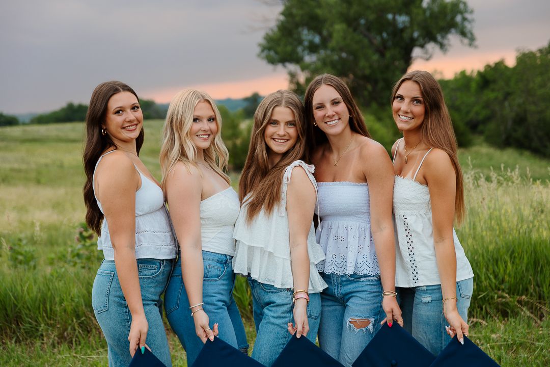 Five young women in white tops and jeans smiling together in a green field at sunset.