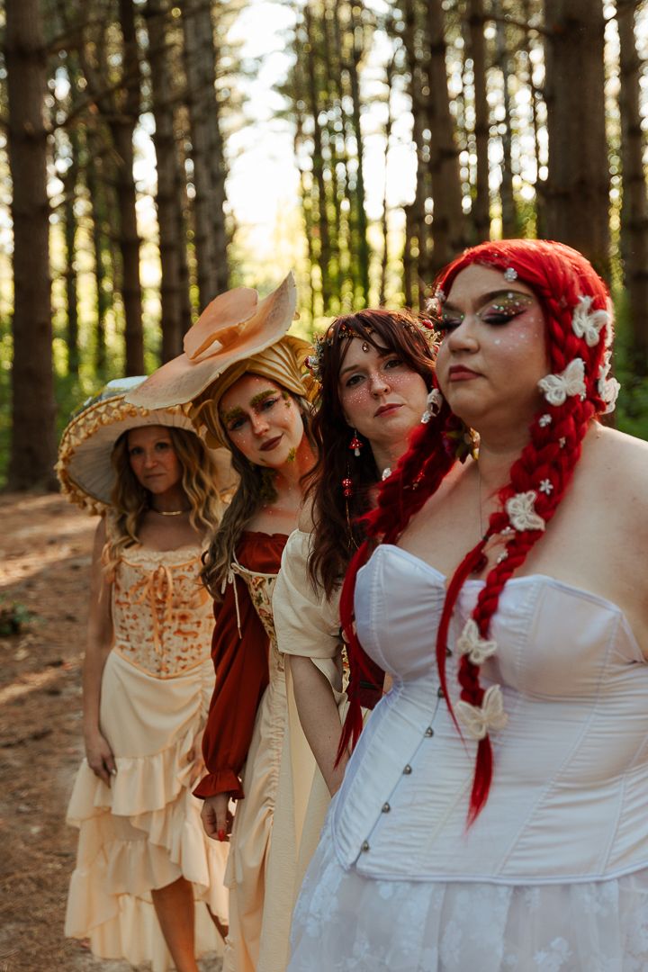 Four women in fantasy costumes pose in a sunlit forest, featuring red hair, floral accents, and vintage-inspired dresses.
