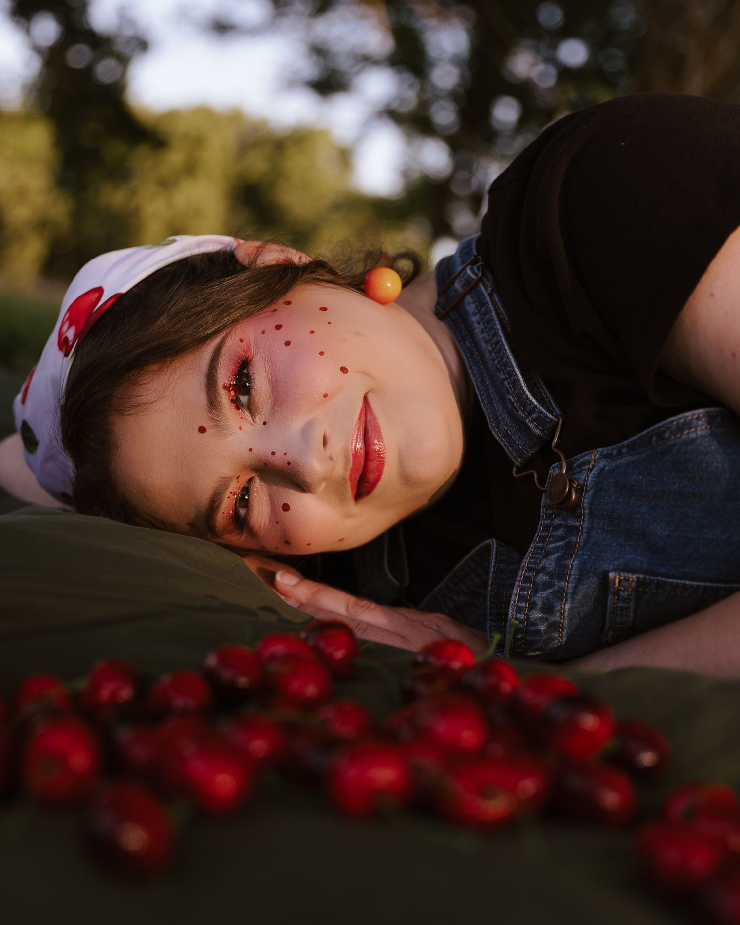 Young woman with cherry-themed makeup and freckles lying near red cherries outdoors.