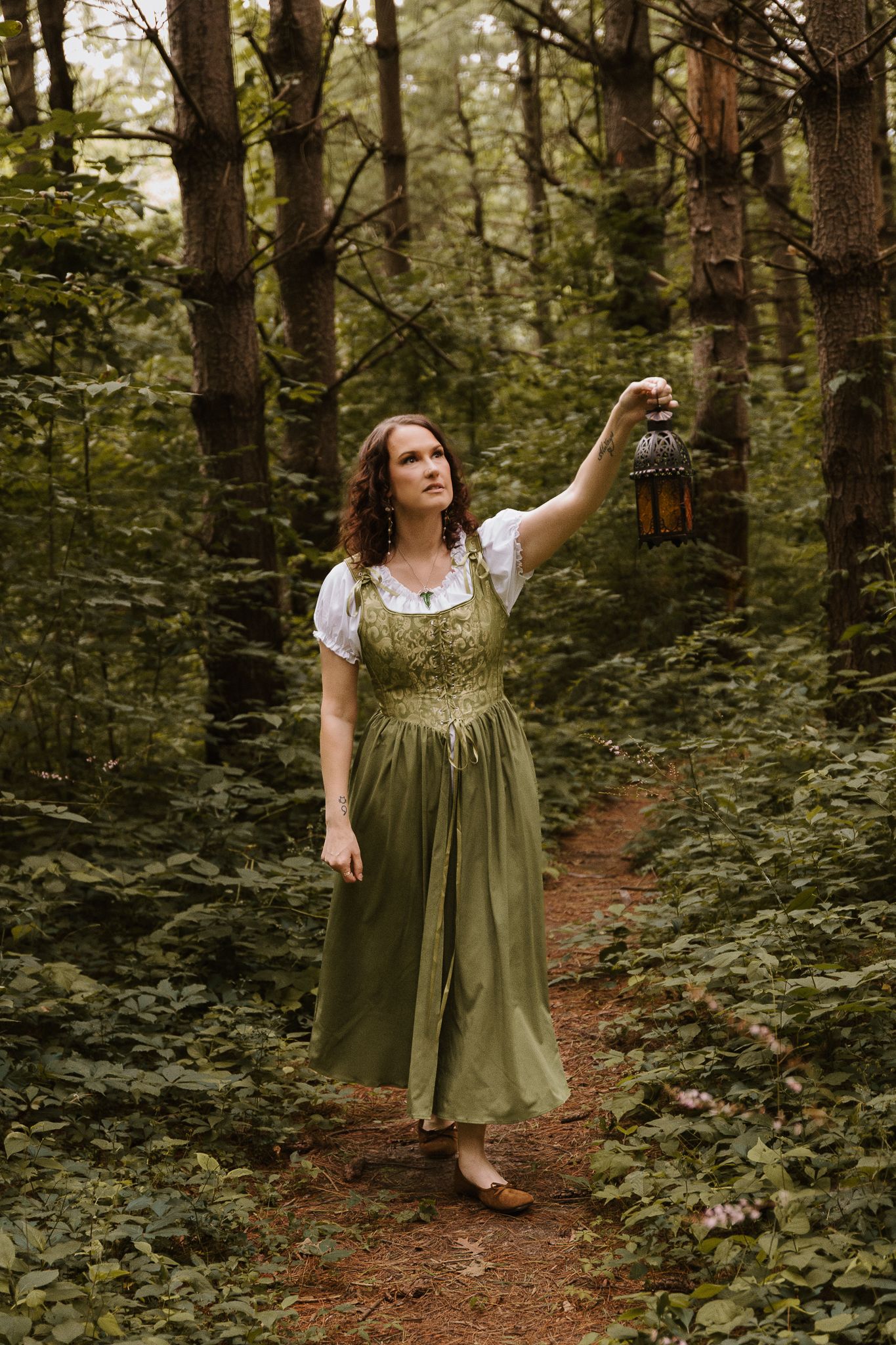 Woman in green medieval dress holding a lantern on a forest path surrounded by lush greenery.