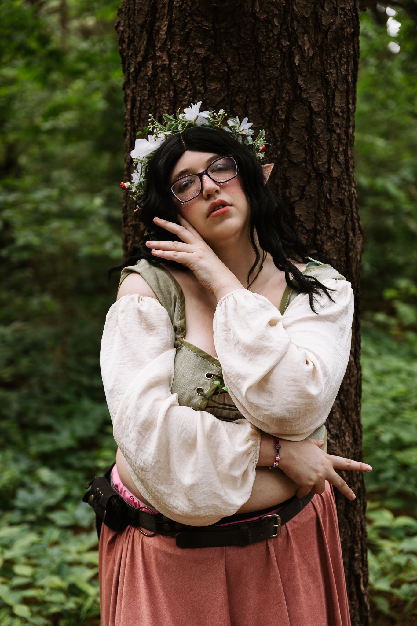 A woman in elf cosplay with pointed ears, flower crown, glasses, and medieval costume leans against a tree in a forest.