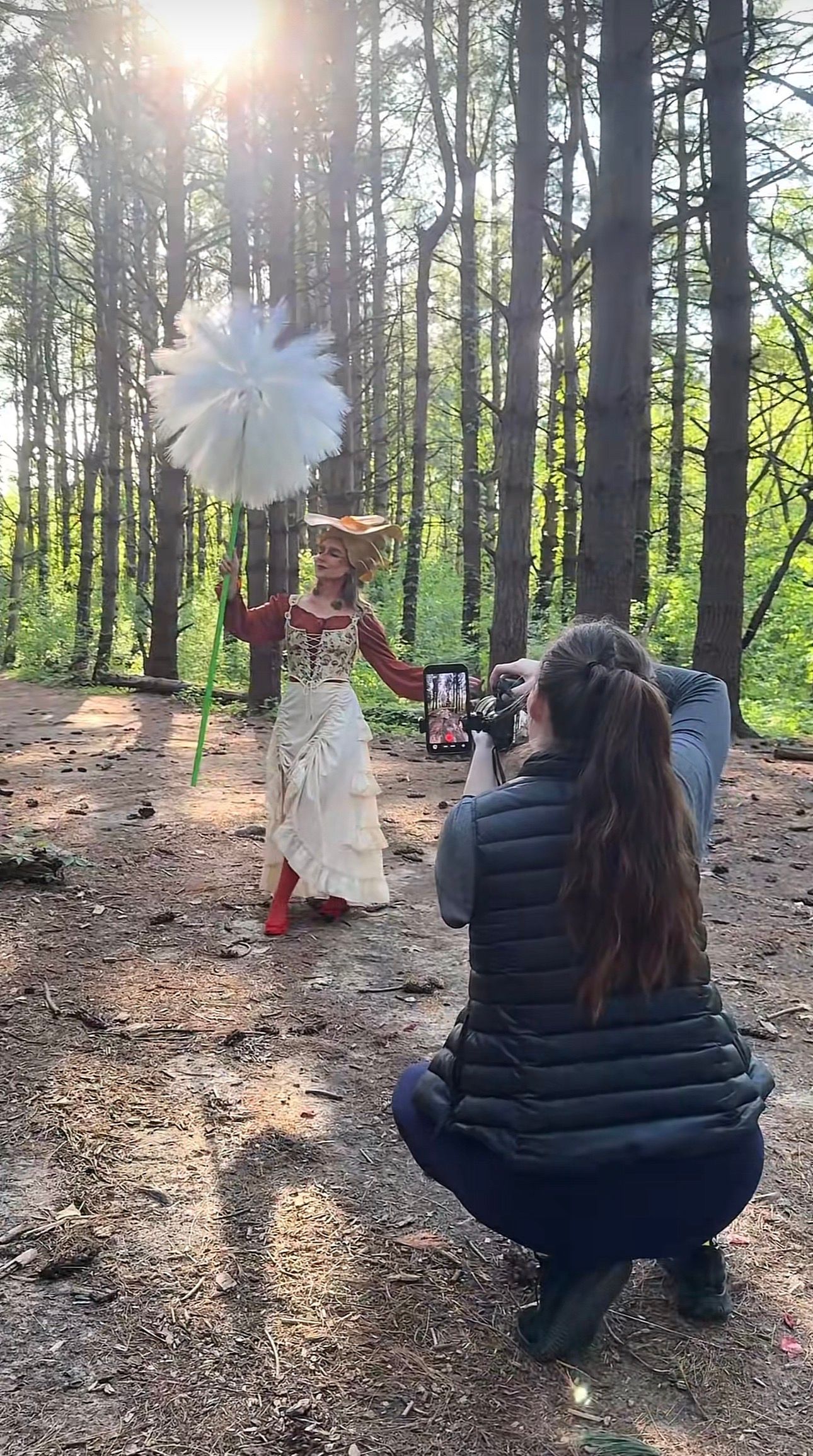 Woman in elaborate costume with feathered fan poses in sunlit forest while photographer crouches to capture shot on smartphone.