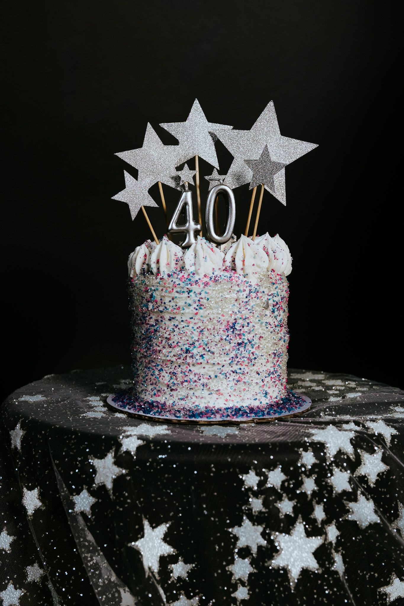 A glittery 40th birthday cake covered in colorful sprinkles, topped with silver star decorations on a dark background.