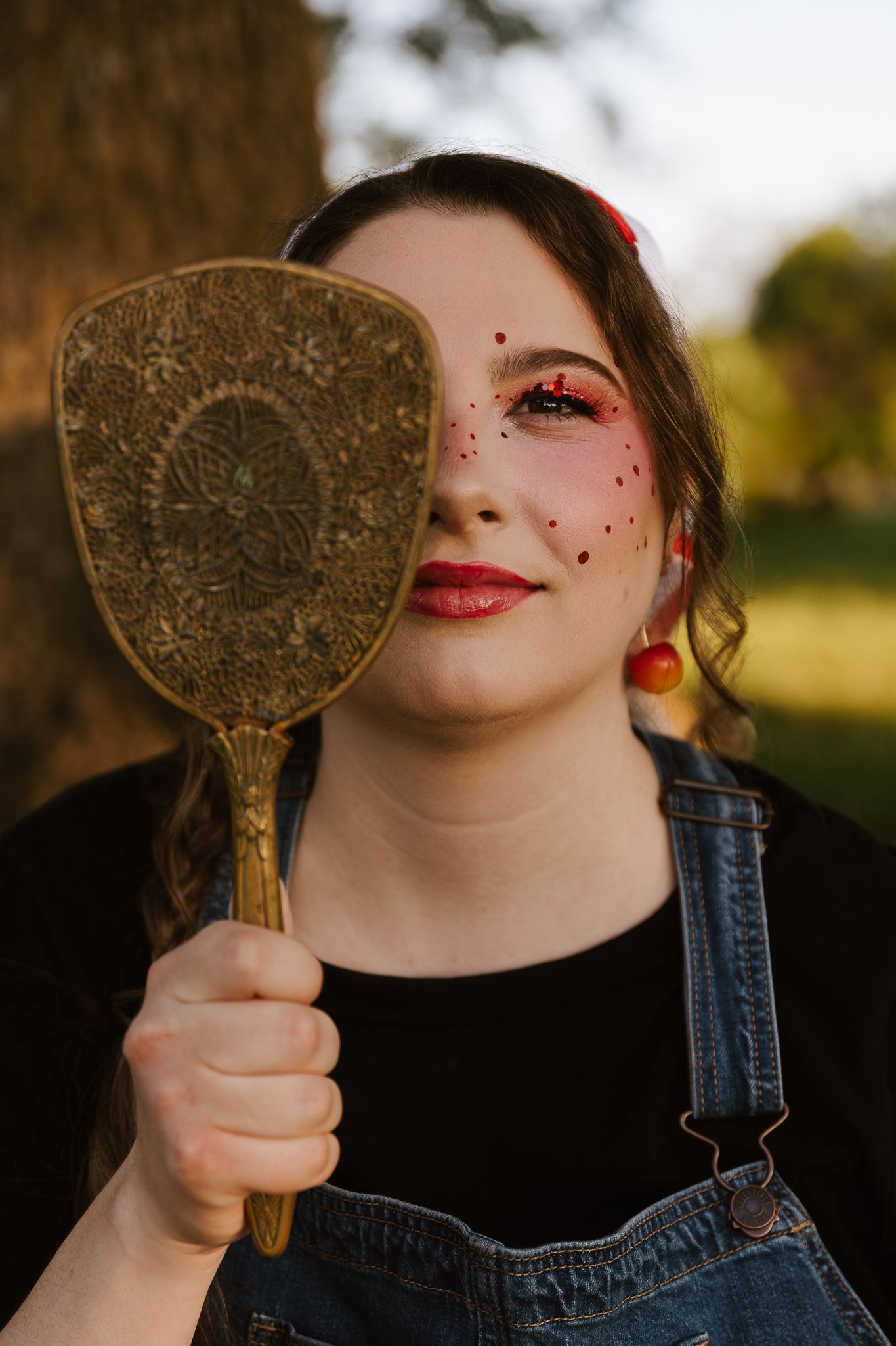 Young woman in denim overalls holding a vintage hand mirror over one eye with decorative face makeup.