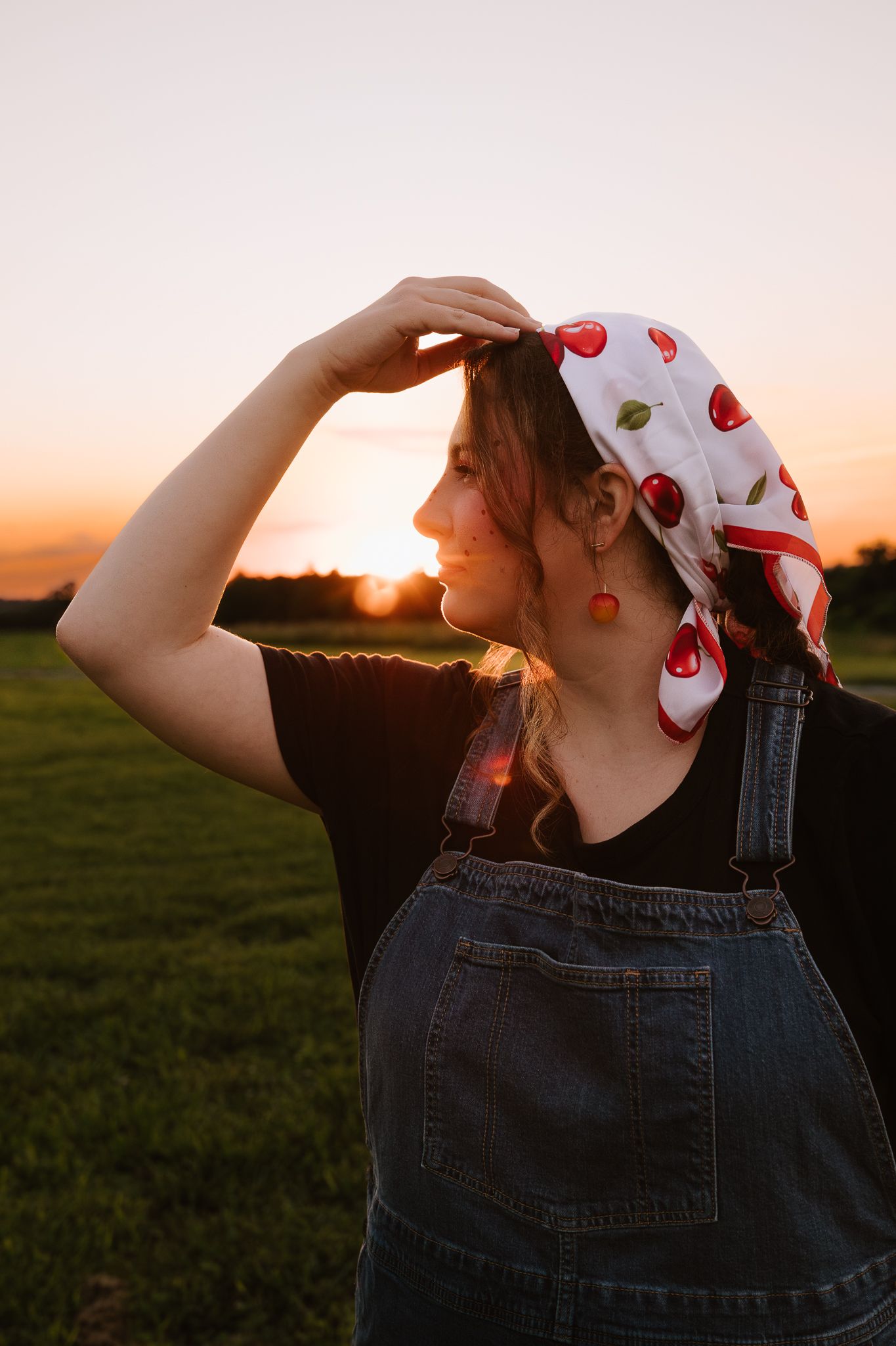 Young woman in denim overalls and cherry print headscarf shielding eyes from golden sunset in green field.