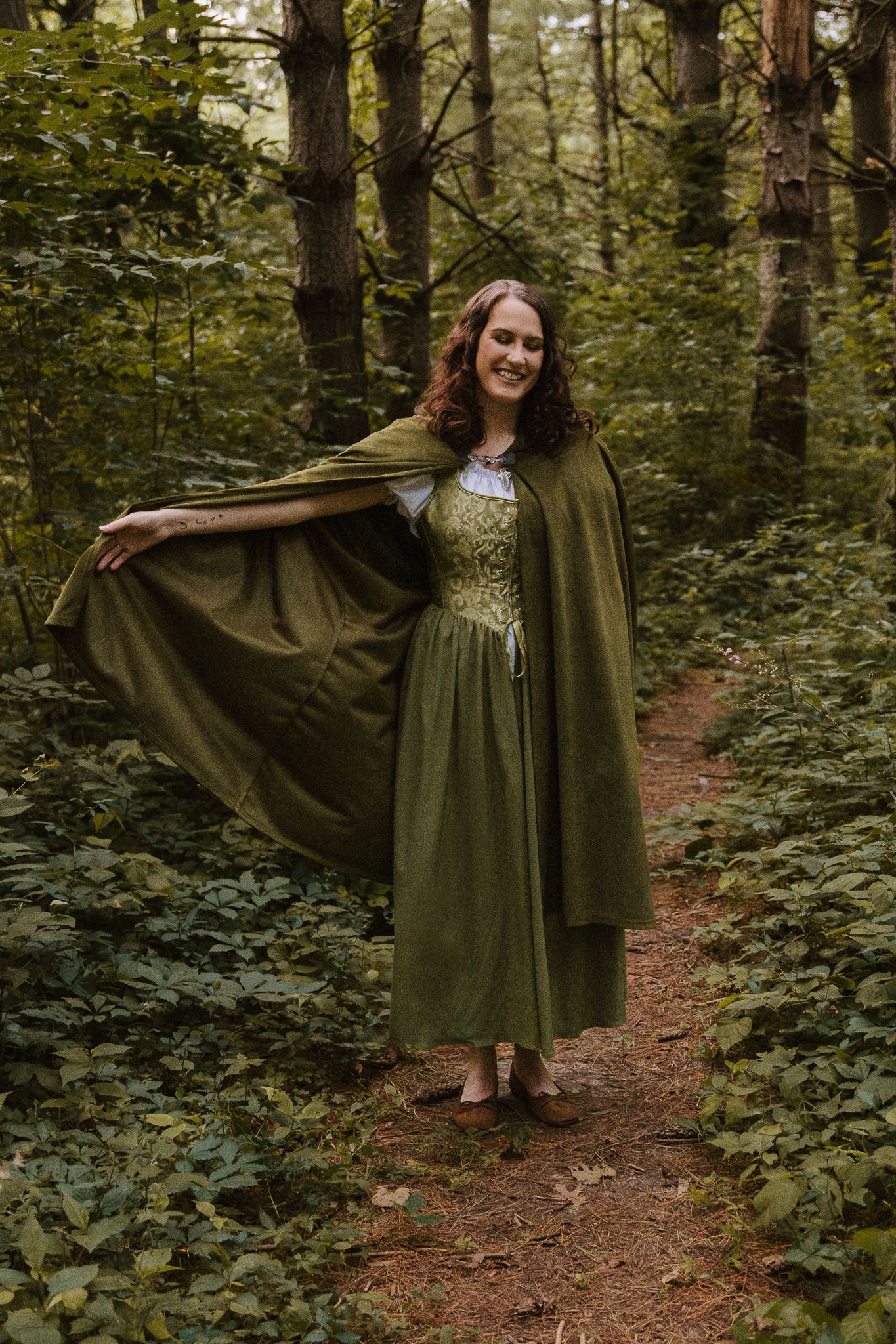 Woman in green cloak and medieval dress smiling on a forest path.