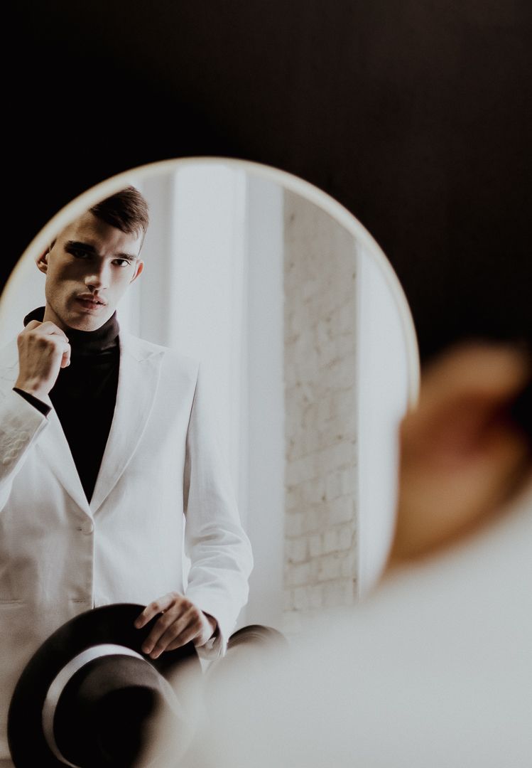 Figure in crisp white suit and black turtleneck poses in moody studio setting.
