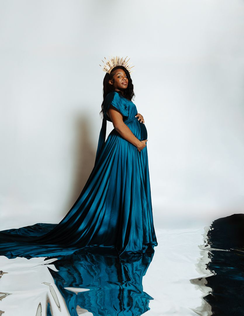 Pregnant woman in teal maternity gown with gold crown poses gracefully in studio.