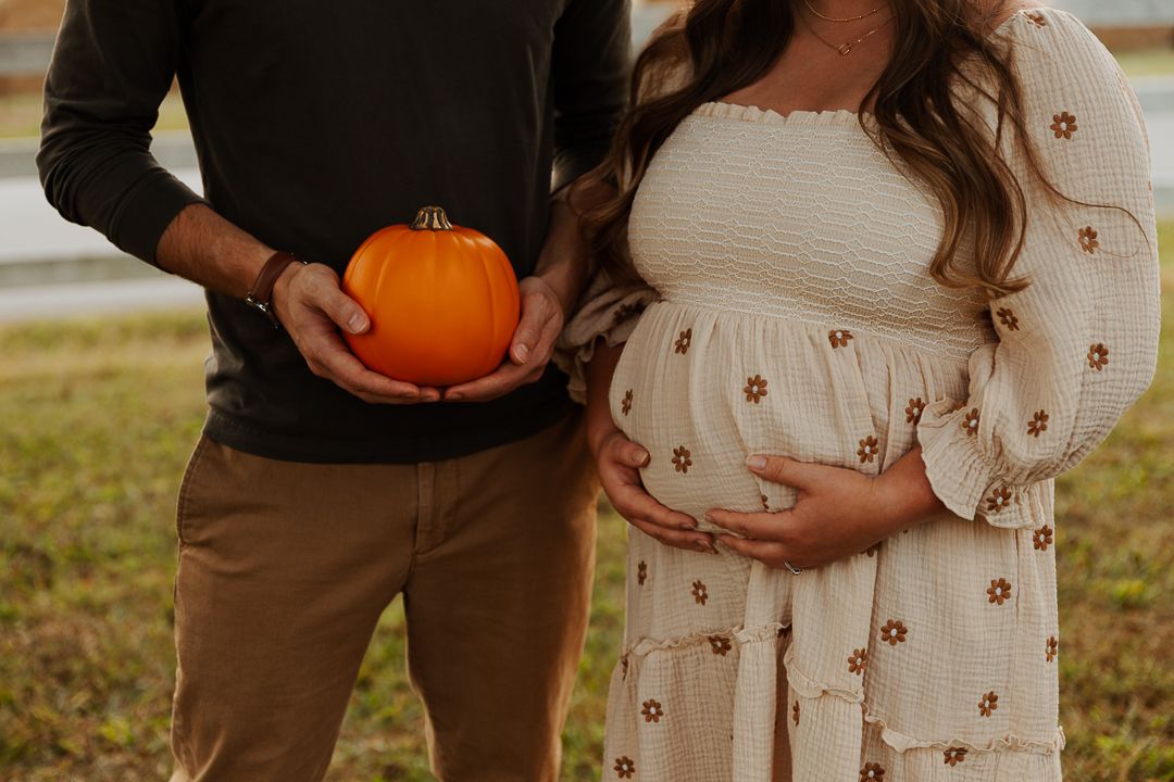 A couple holding a pumpkin on a fall day outdoors in a casual outfit stands close together.