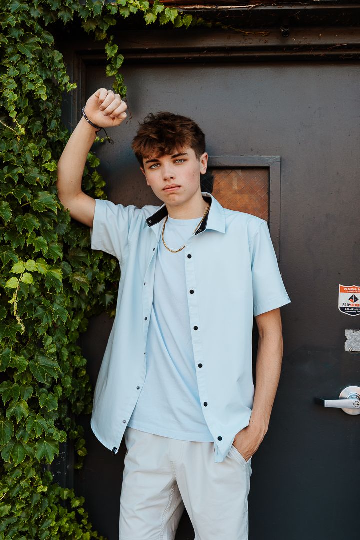 Person in light blue attire poses against dark doorway with greenery and brick backdrop.