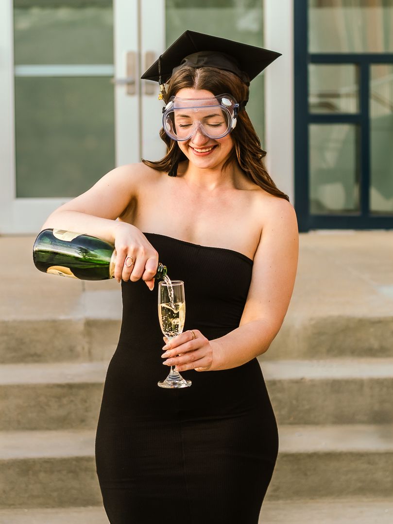 Person in black dress pours champagne into a flute glass while seated on outdoor steps in warm sunlight.