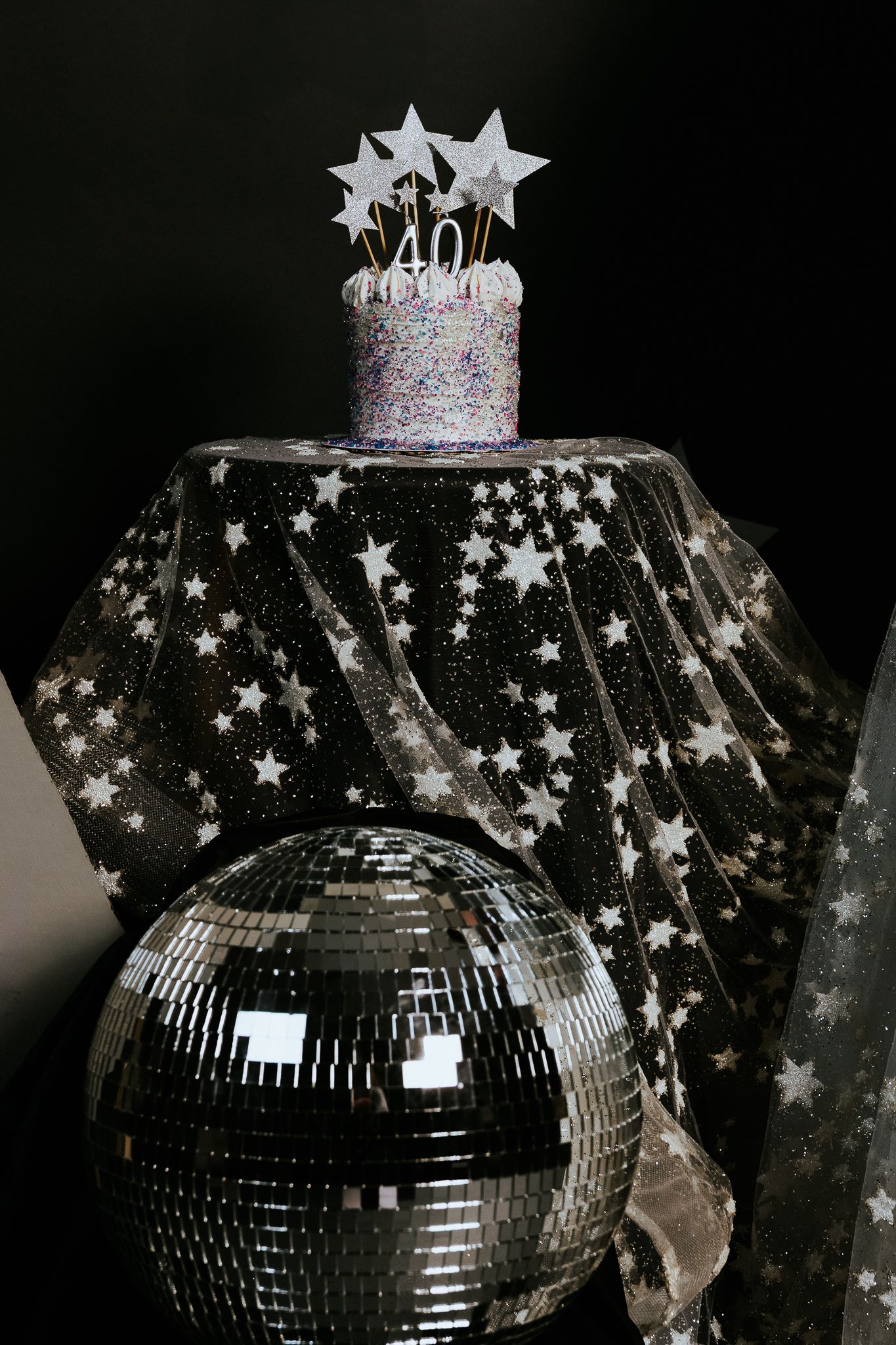 Glittery 40th birthday cake with star toppers on a star-printed cloth beside a disco mirror ball.