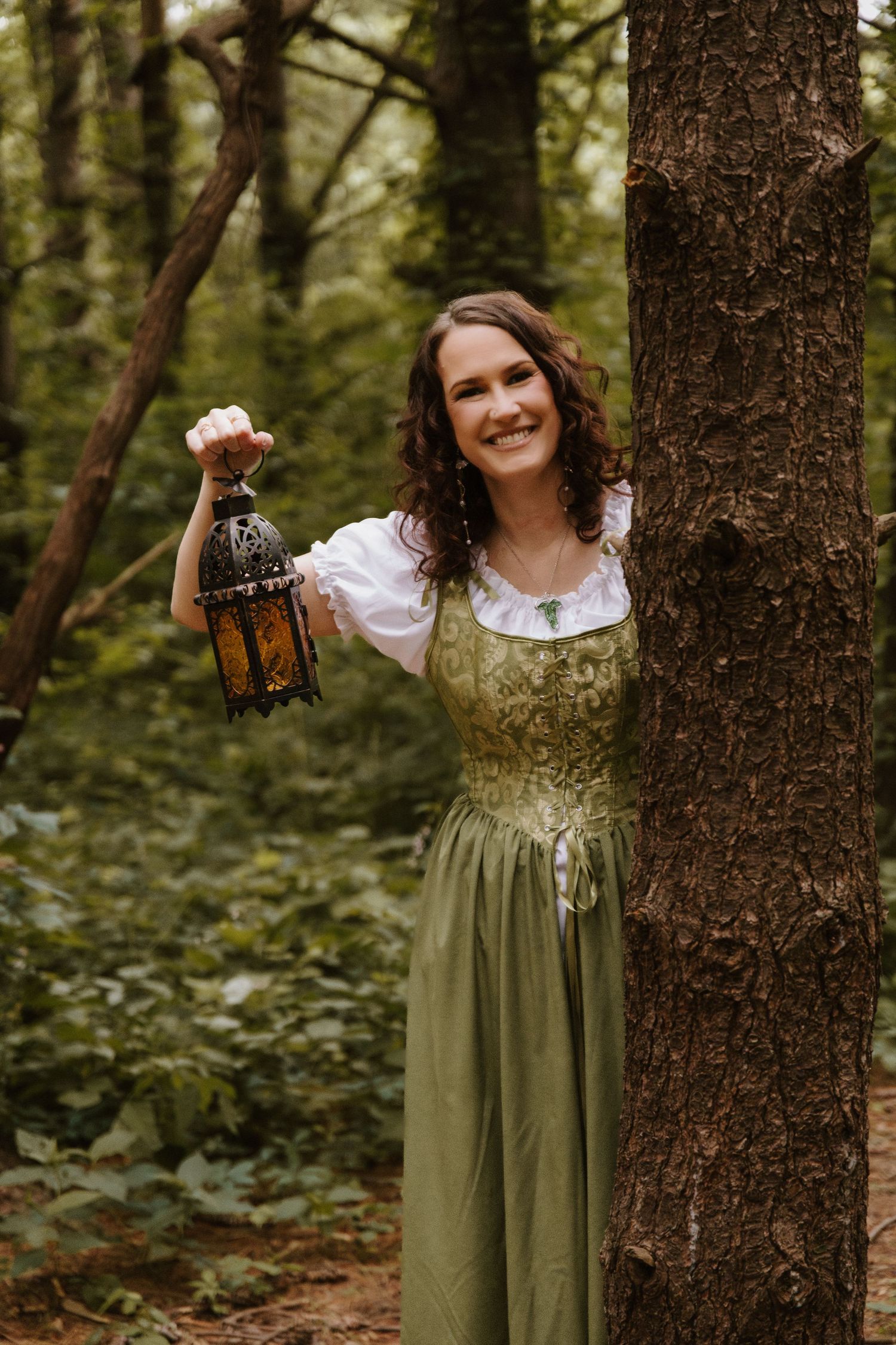 A smiling woman in a green medieval dress holds a decorative lantern while posing beside a tree in a lush forest.