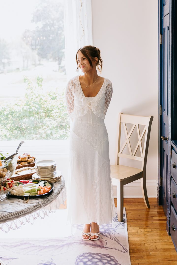 Bride in white lace long-sleeve gown standing by window near food table with white sandals on floor.