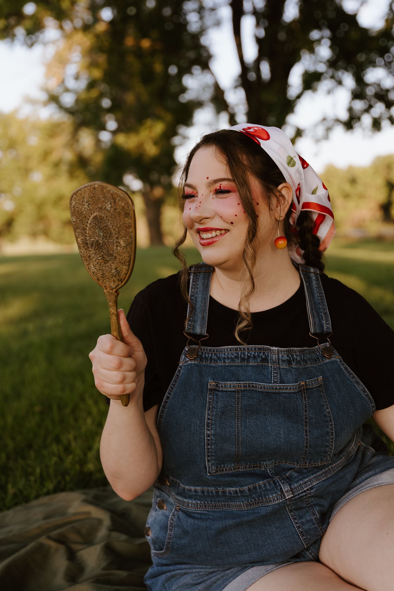 Smiling woman in denim overalls and cherry bandana holds vintage mirror while sitting outdoors in park.
