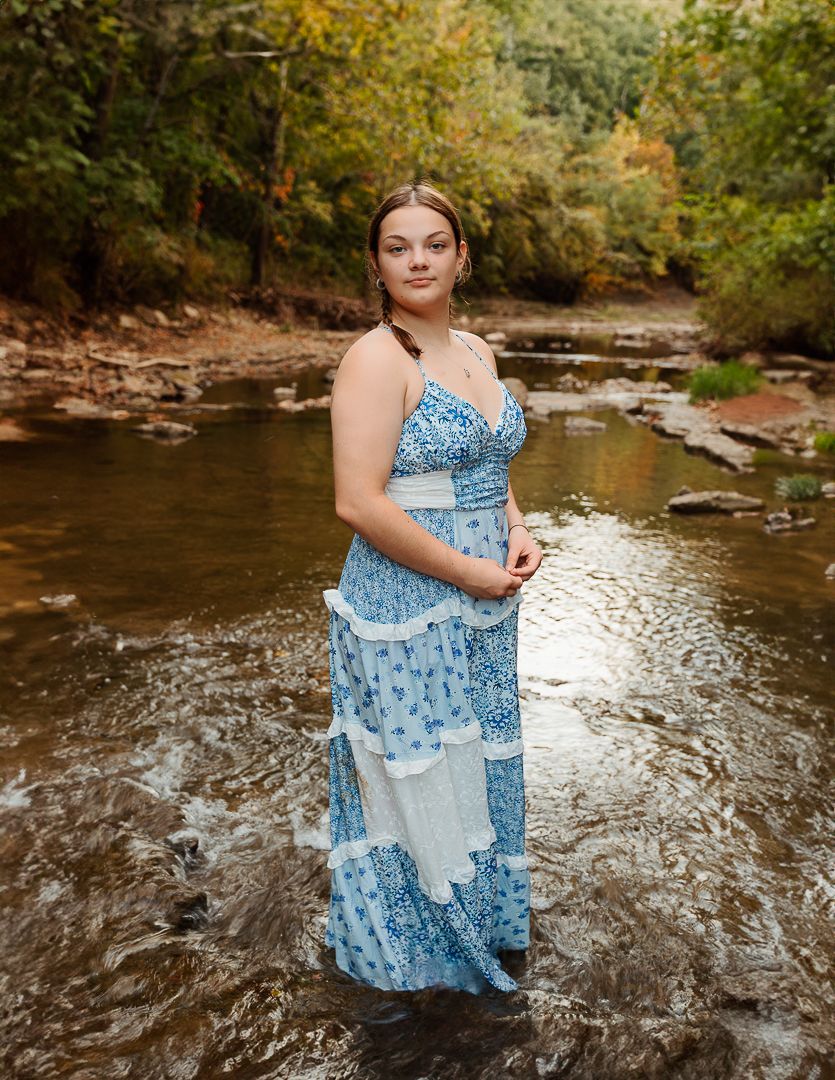 A woman in a flowing blue dress stands in a shallow creek with rocks and autumn foliage surrounding the scene.