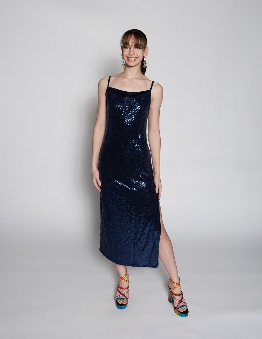 Woman wearing a navy blue sequin midi slip dress with spaghetti straps and a side slit, paired with colorful heeled sandals.