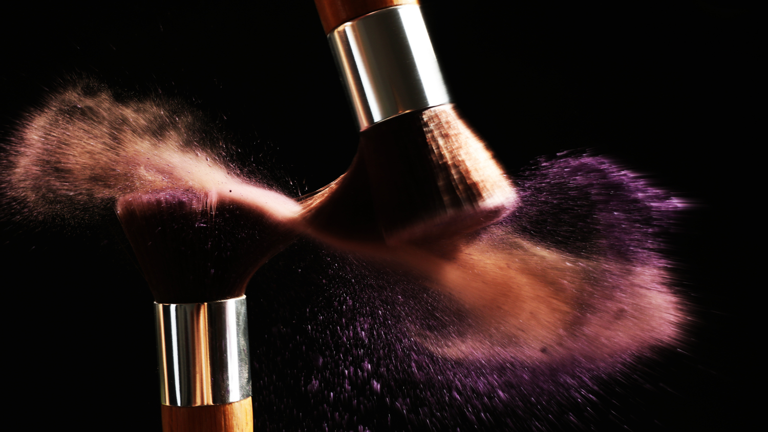 Two makeup brushes colliding with colorful pink and purple powder explosion on black background.