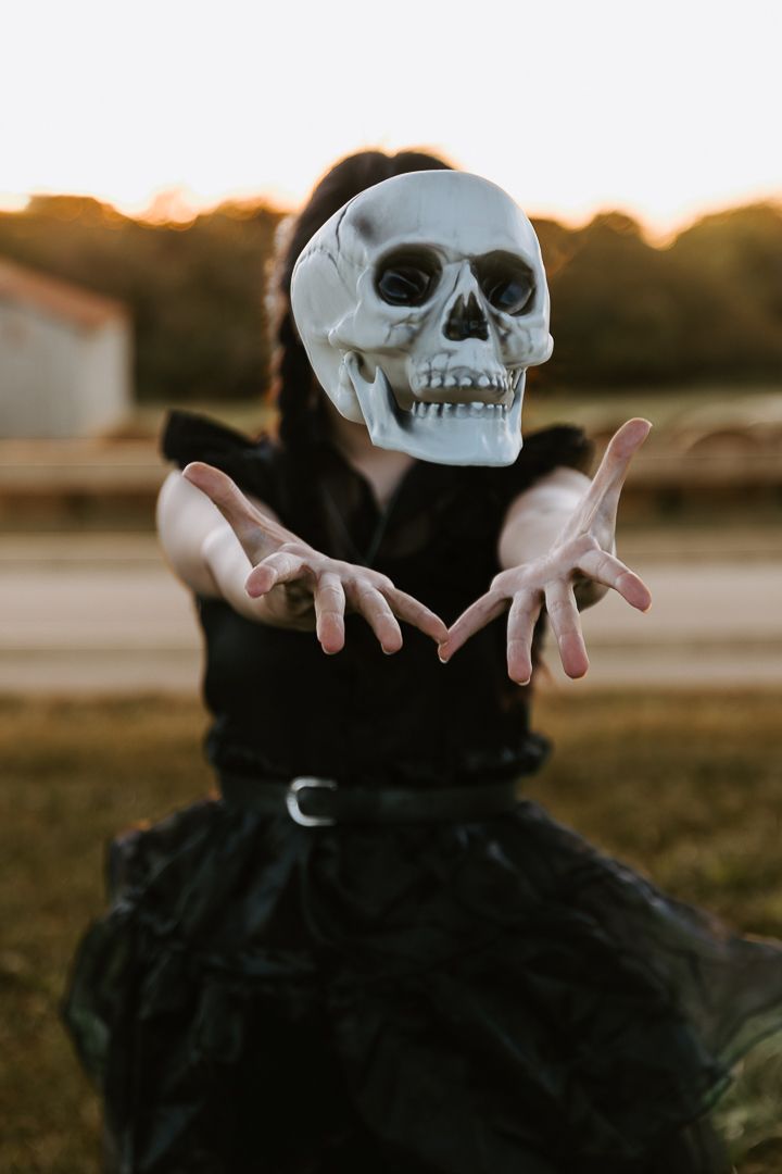 A person wearing black clothing holds up a white skull mask against a sunset backdrop in a spooky Halloween pose.