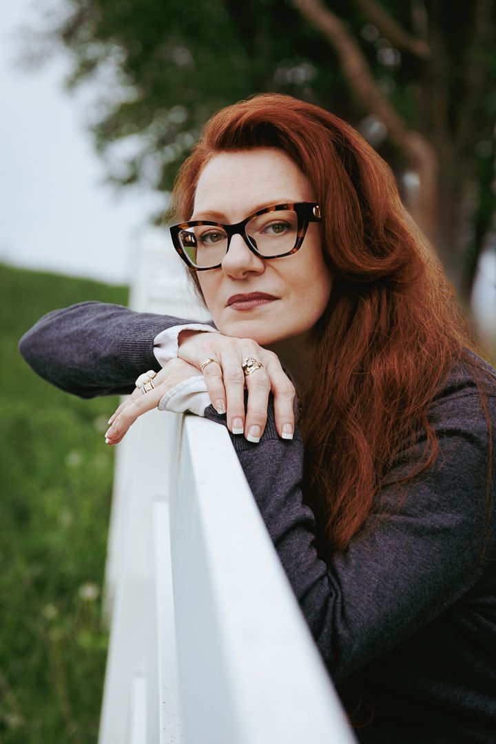 A person with red hair and glasses leans on a white railing outdoors in a contemplative pose.