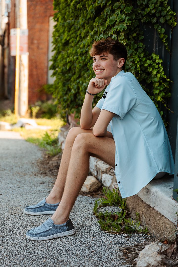 A casual portrait of someone in a light blue shirt sitting on concrete steps next to ivy-covered walls.